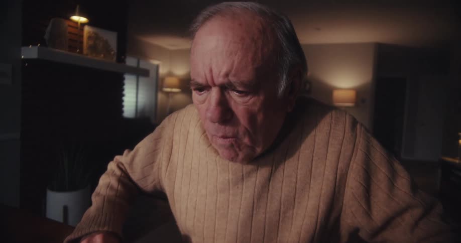 An elderly man sits at a table in a dimly lit living room his face suddenly showing surprise as he works on his laptop