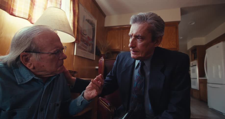 An elderly man talks with a businessman who is presenting a financial chart on a tablet in a home setting
