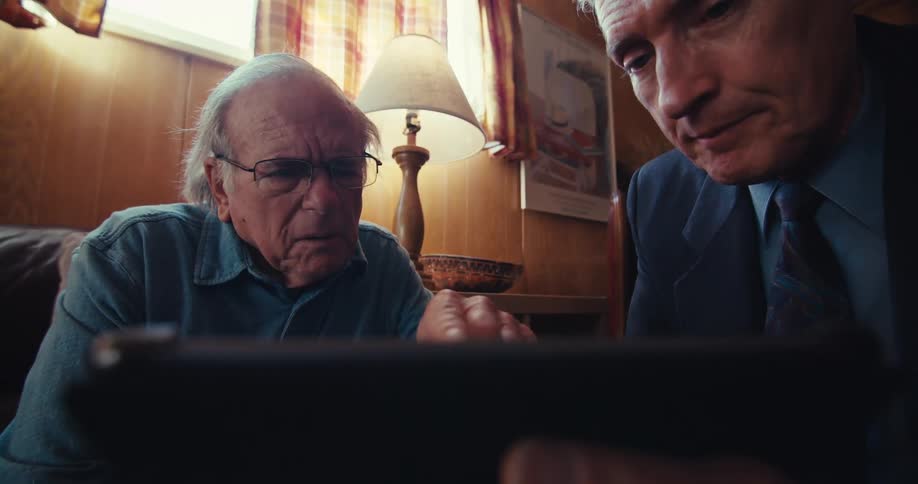 An elderly man and a middleaged man focus on a tablet in a warmly lit room
