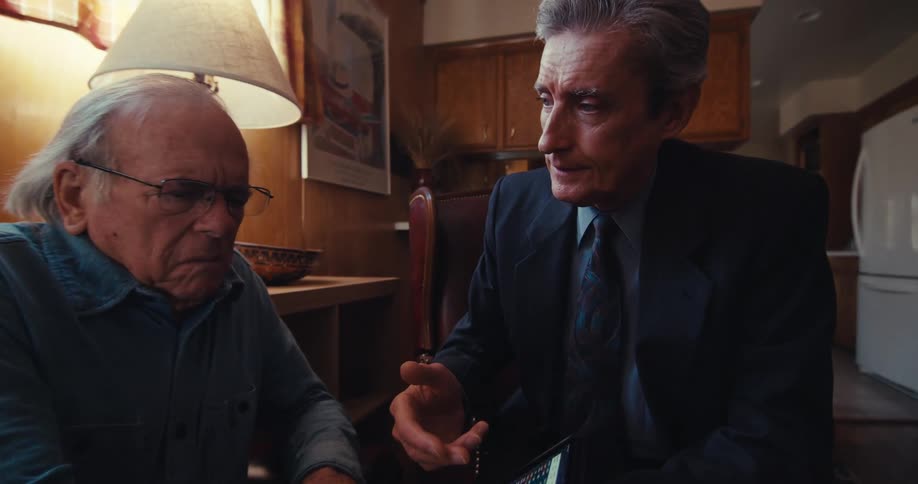 An advisor in a suit shows an elderly man seated beside him on a couch something on a tablet pointing to the screen while explaining