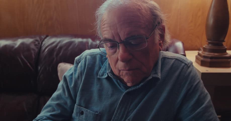 An elderly man gestures toward a trading chart presented by a man sitting beside him in a home setting