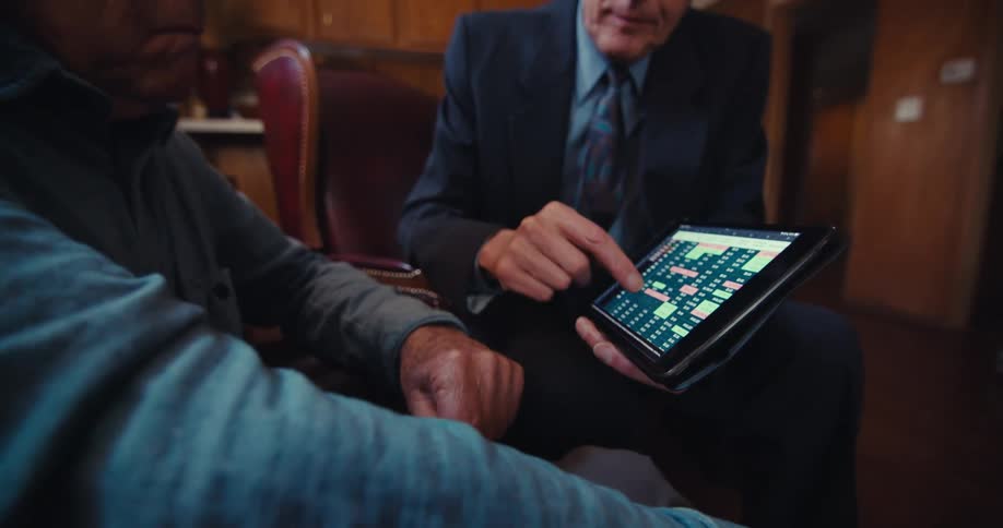 A man in a suit is engaged in a discussion showing an elderly man a tablet with a chart displayed on the screen