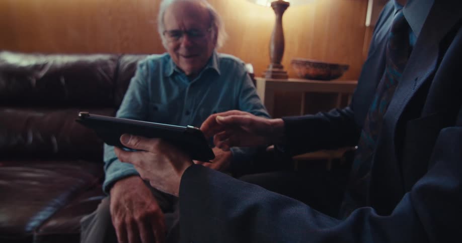 An elderly man shakes hands with a businessman who is presenting information on a tablet in a home setting