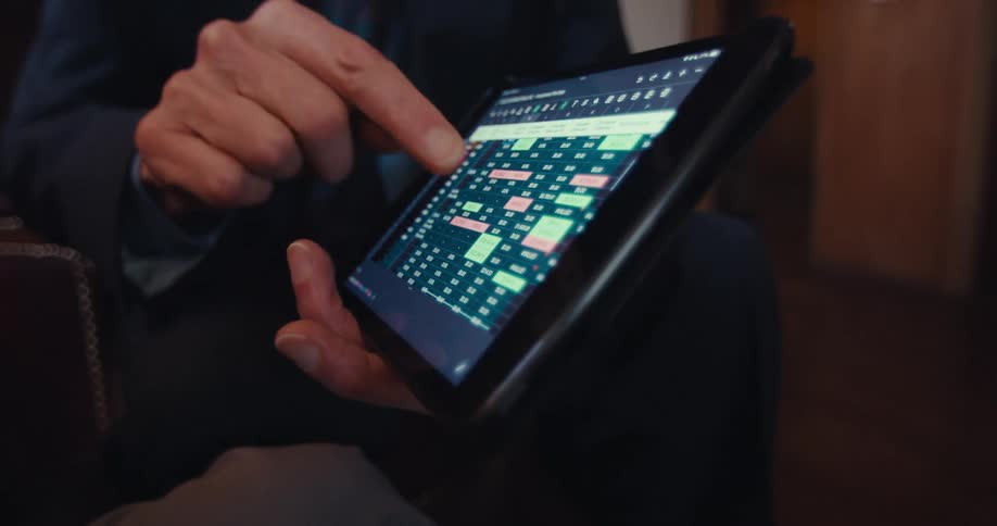 A person wearing a suit holds a tablet using their finger to point at and interact with a financial chart or spreadsheet displayed on the screen