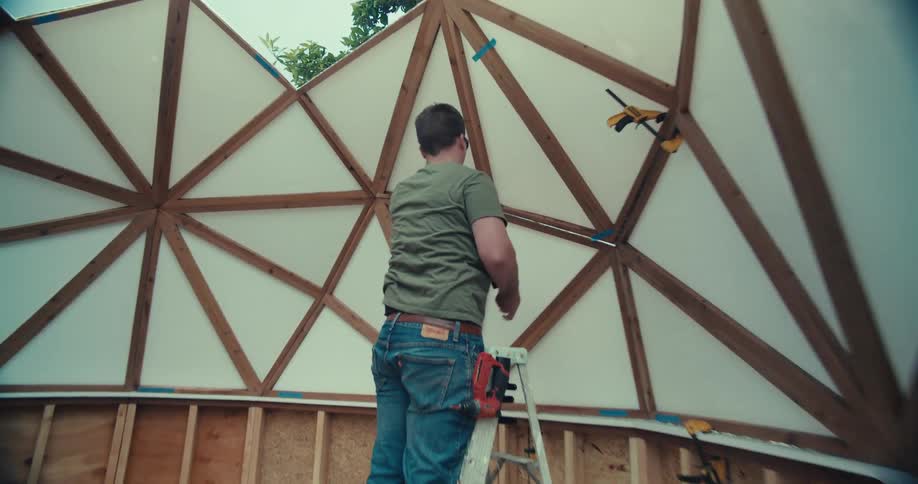 A man stands on a ladder in a backyard actively constructing the wooden frame and attaching panels to a geodesic dome structure he is building