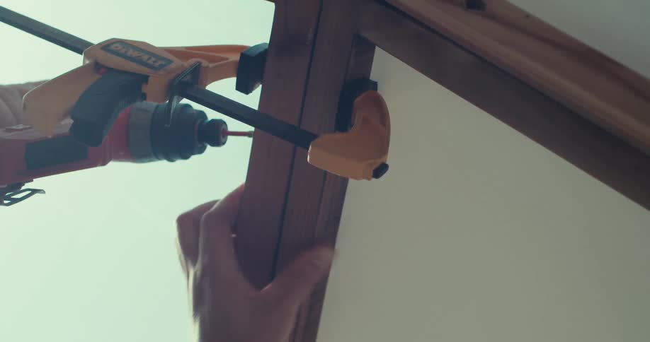 A person drills a nail to join two pieces of wood during the construction of a shelter at midday