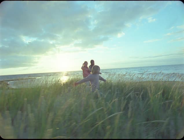 Three children scale a sand dune covered in tall grass on a beach with the setting sun casting a warm glow beneath the cloudy sky