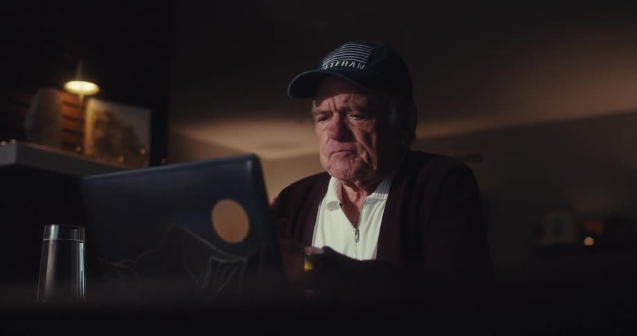 An elderly man wearing a veteran cap sits in a dimly lit room focused intently on the screen of his laptop