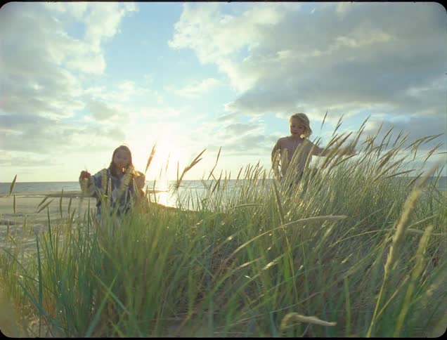 Three children scale a sand dune covered in tall grass on a beach with the setting sun casting a warm glow beneath the cloudy sky