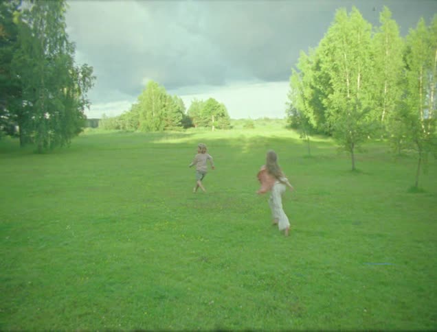 Children are running across a lush green field with trees in the background under a cloudy sky