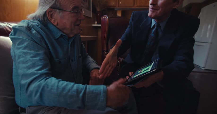 Older businessman explains to an elderly customer using a tablet while sitting in the living room and they make a deal