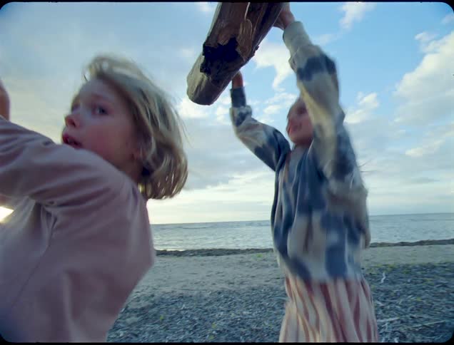 Three children hang from a large weathered tree branch suspended over a rocky beach near the ocean at dusk