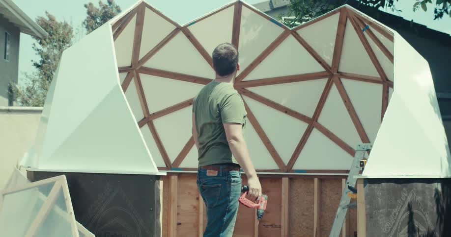 A man is building a shed in his backyard on a sunny day wearing sunglasses holding a drill and stepping off a ladder to observe his construction progress