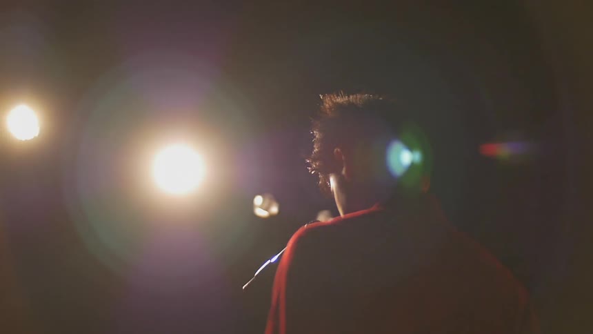 A teenage boy stands in front of a microphone under bright lights