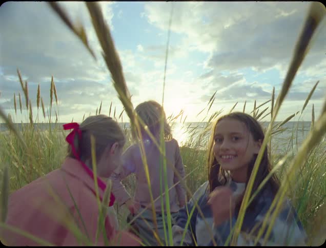 Three children sit amongst tall grass on a beach under a cloudy sky with the sun setting in the distance