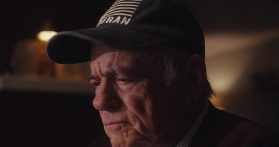 An elderly man wearing a baseball cap looks sad in his living room
