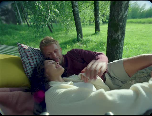 A woman in white clothes is walking up to a man who is lying in a hammock hanging from two trees and she lays him down with him