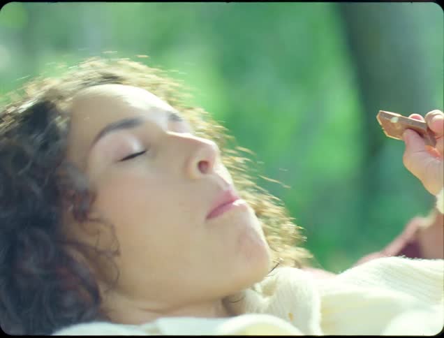 A couple of young lovers sitting in a hammock in the park while she is enjoying a piece of peanut chocolate