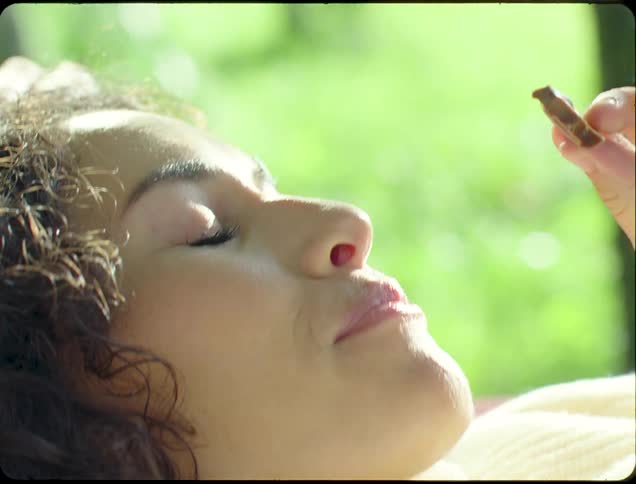 A person is lying down outdoors wearing a white sweater eating a candy bar surrounded by green foliage in the background