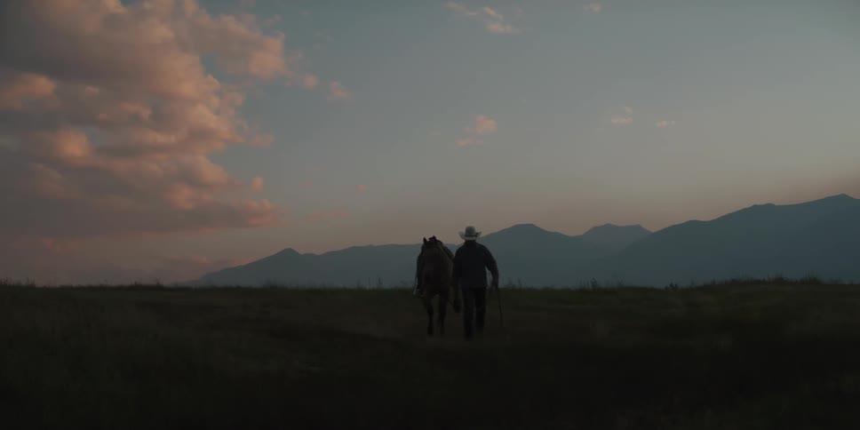 A cowboy walks alongside his horse through a vast dimly lit grassy field towards the camera silhouetted against a backdrop of mountains and a dusky sky
