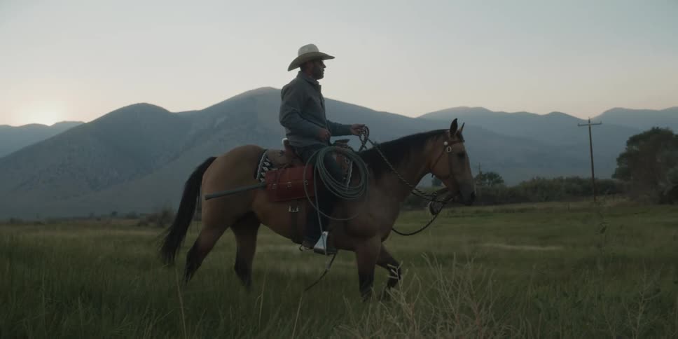 A man in a cowboy hat rides a horse through a field with distant mountains silhouetted against the soft light of the morning sky