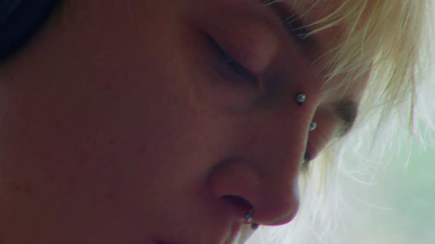 A woman with a nose piercing holds a pen and writes while sitting beside a window