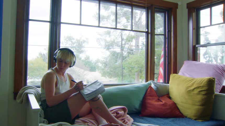 A man relaxes by a sunlit window writing in a notebook and nodding to music with headphones on