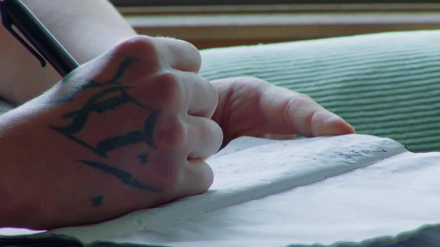 A tattooed hand writes in a notebook with a pen