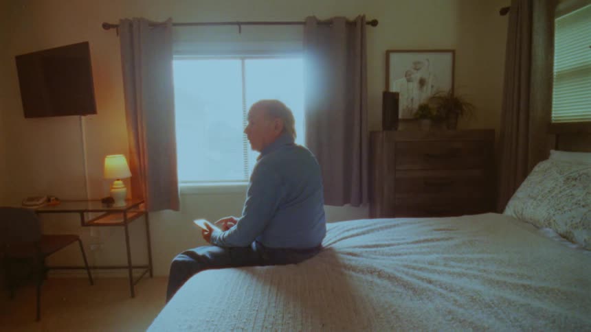 An elderly man sits sideways on the edge of his bed holding a mobile phone and gazing towards the brightly lit window in his bedroom during the day