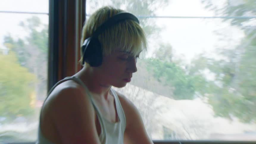 A short blond hair woman wearing headphones sits focused near a window writing or drawing intently in a notebook with a pen