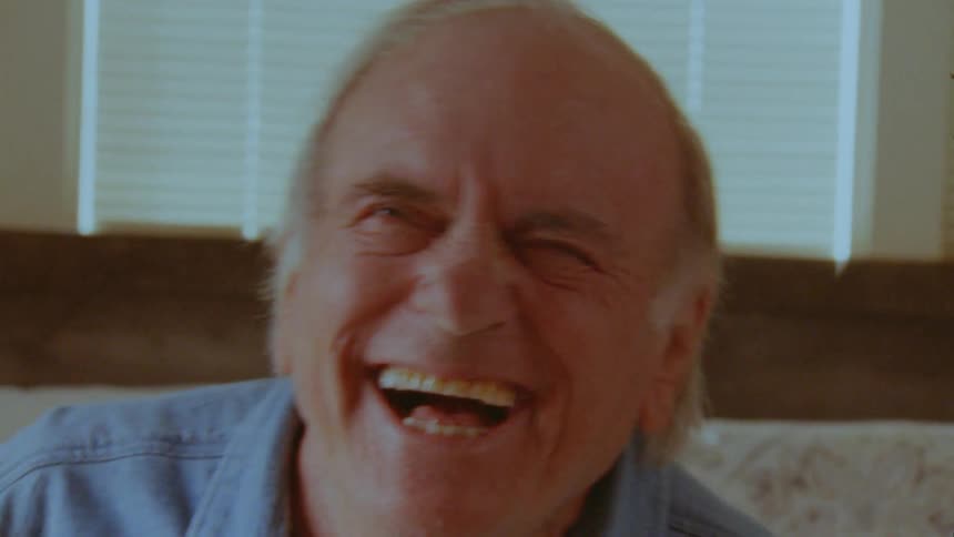 An elderly man sitting indoors appears to be talking to someone before breaking into hearty laughter