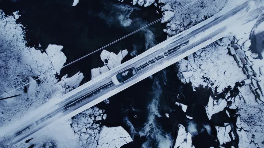 An aerial drone shot follows a single car as it drives across a snowcovered bridge spanning a dark river filled with chunks of ice in winter