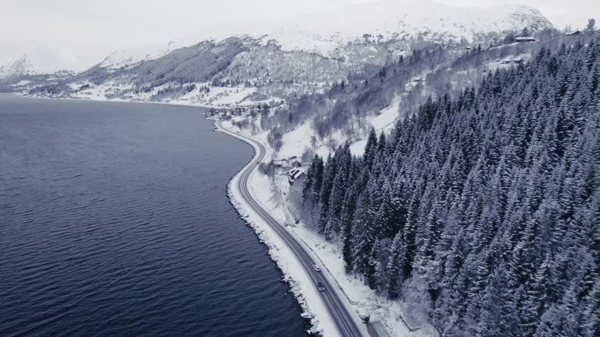 An aerial shot follows vehicles driving on a road that winds alongside a large water body bordered by a snowcovered forest and mountains