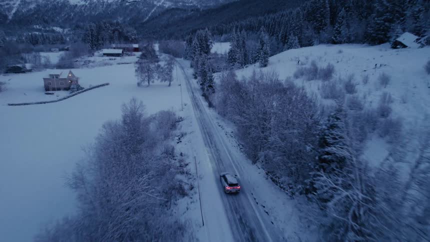 The car drives in the snow past the homes near the mountains