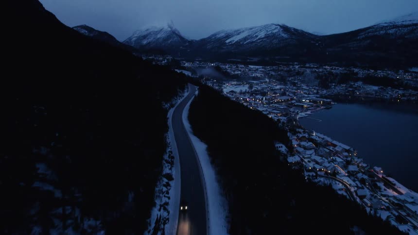 An aerial view at dusk follows a car with headlights driving on a snowlined road beside a lake revealing a lit coastal town nestled among snowy mountains