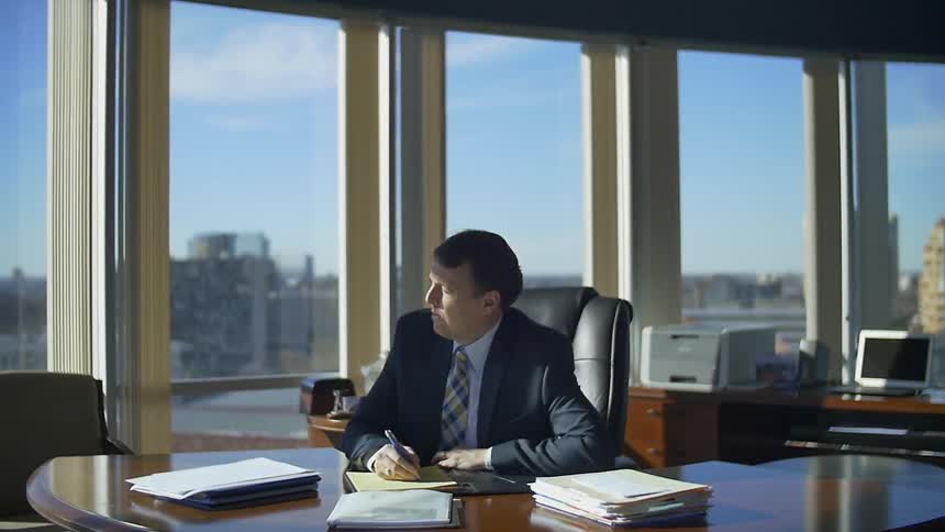 Business man writes on a yellow legal pad then looks away in deep thought