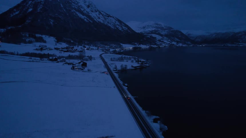 An aerial view at dusk captures a car with its headlights on driving along a road through a snowy landscape beside a dark body of water with mountains and distant settlement lights