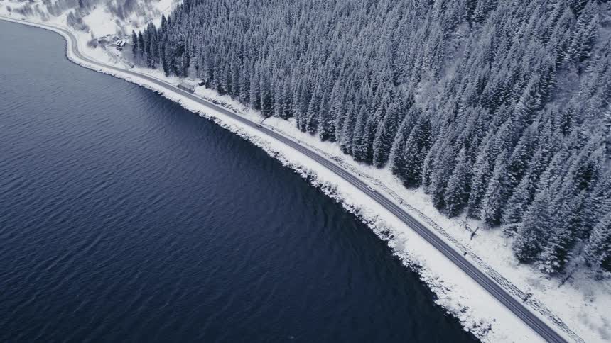 An aerial shot tracks cars driving along a road bordered by a dark rippling body of water on one side and a dense snowcovered pine forest on the other