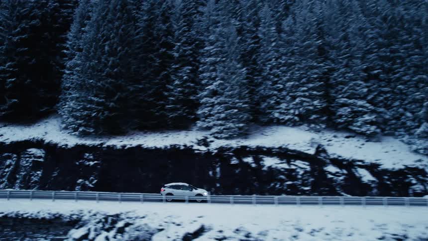 A white car partially covered in snow drives along a road as the camera zooms out to reveal a vast snowy landscape with a dense pine forest and a body of water