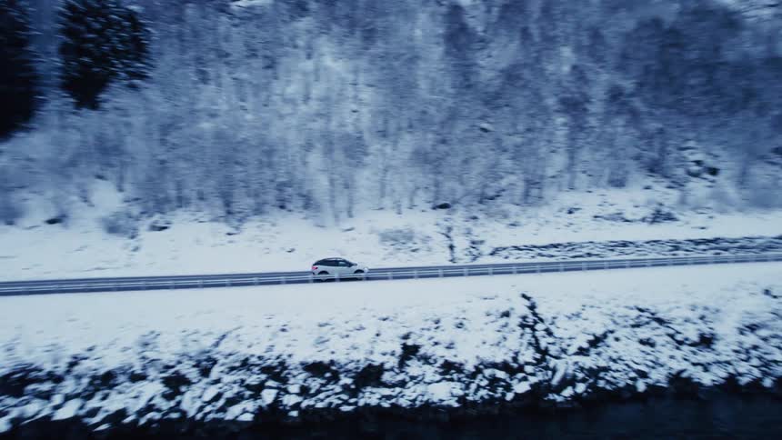 An aerial shot follows a car driving along a snowlined road that curves beside a large body of water with steep snowcovered mountains rising on either side