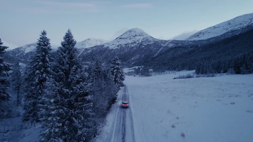 An aerial shot lowers and tracks a car with its taillights on as it drives along a snowcovered road through a valley flanked by snowladen forests and mountains under a dim sky