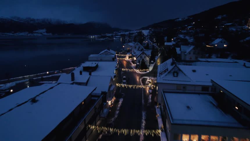 An aerial view at night shows a snowcovered coastal town with streets illuminated by strings of lights nestled between a dark water body and mountains
