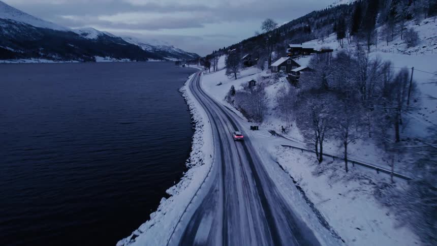 An aerial shot descends to follow a car driving along a snowcovered road at dusk with a dark body of water on one side and snowcovered houses and mountains on the other