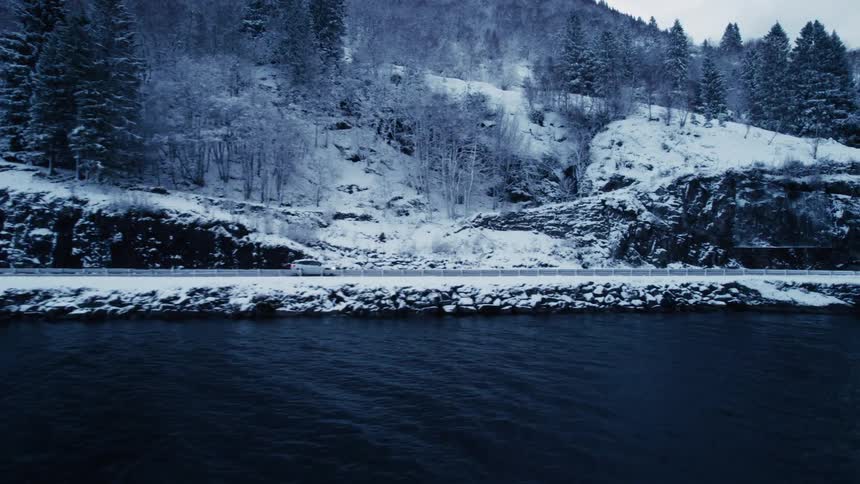 A white car drives along a snowlined road that runs parallel to a dark body of water with a snowcovered forested mountainside rising above