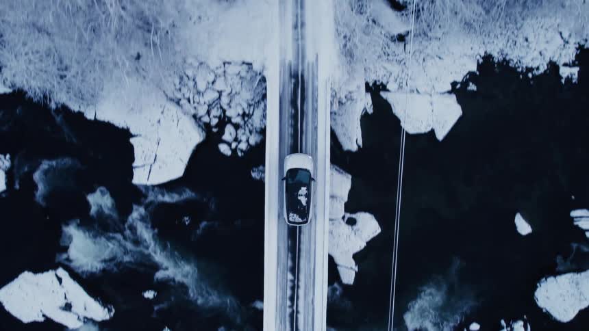 A topdown aerial view follows a car driving across a snowcovered bridge over an icy river then continues along a road through a snowy treelined landscape