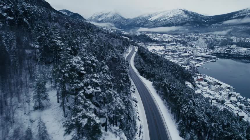 An aerial shot flies down the slope of a snowcovered forested hill revealing a road leading to a coastal town nestled by a body of water with mountains in the distance