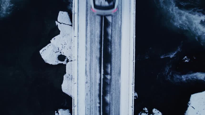 A white car driving on a small bridge that passes over a river with a strong current of water seen from above