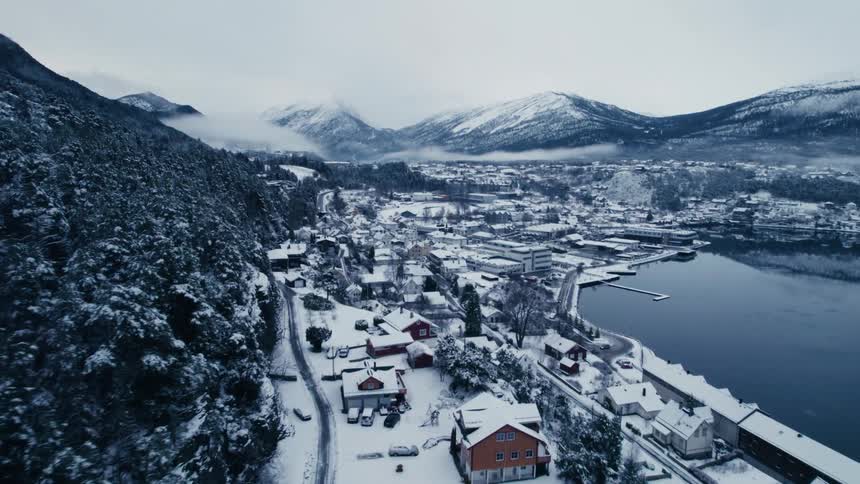 An aerial shot flies over a snowcovered coastal town nestled between a forested slope and a calm body of water with larger mountains in the background