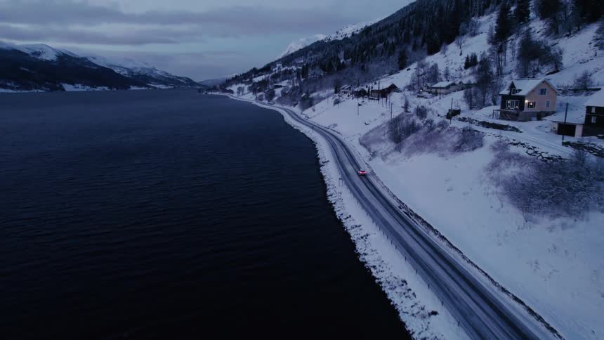 An aerial shot flies down tracking a car as it drives along a snowcovered road beside a dark body of water with houses and mountains visible in the dusky landscape