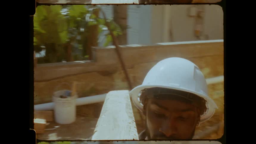 A construction worker in a hard hat carries a wooden beam at a work site with palm trees visible in the background